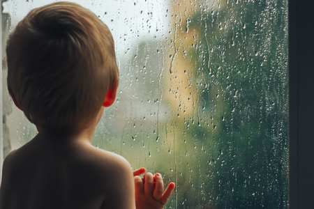 Sad little boy looking out the window on rain drops through wet glass autumn bad weather.の写真素材