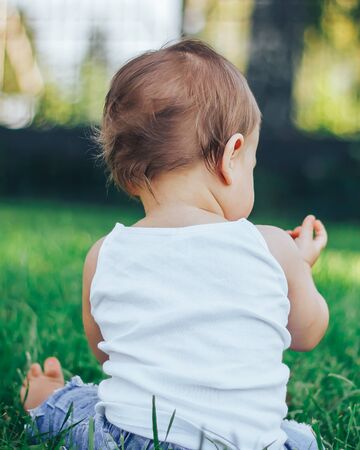 Little girl in white outfit sitting in grass in park looking away from cameraの写真素材