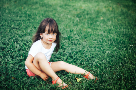 very beautiful little girl sitting on grass with long hair looking at cameraの写真素材