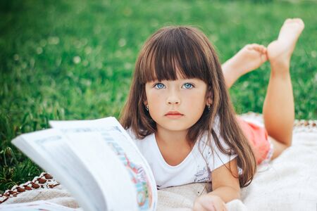 little beautiful girl lie on grass with book looking at cameraの写真素材