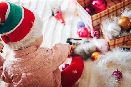 Funny baby boywearing santa claus hat and suit playing with Christmas ball over Christmas tree in room. Holiday season.の写真素材