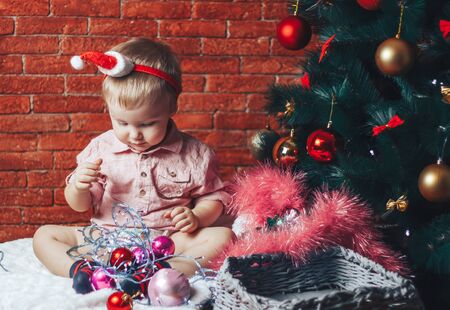 A beautiful baby in a santa hat sits near a Christmas tree with christmas toys and lightsの写真素材