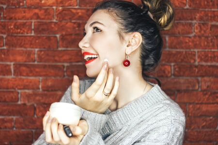 Beauty young smiling girl with white jar of cream looking in the camera and face cream smears on red brick background.の写真素材