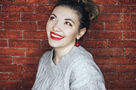 Beautiful woman with red lips, wearing grey sweater, looking up and smiling with teeth, on red brick backgroundの写真素材