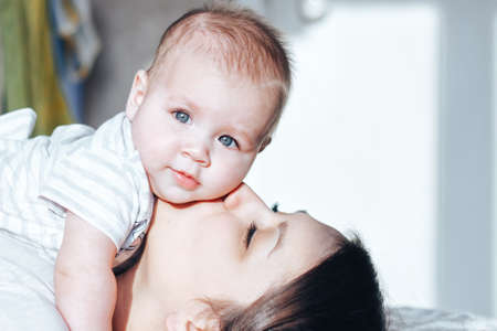 Mum kissing her baby on a white bed. Newborn looking at the camera. Happy motherhood concept,...