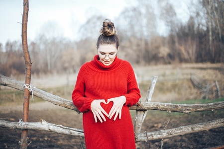 Pregnant woman standing in a field holding his stomach, waiting for the child, summer or spring day, a young mother resting in the open air, in red sweater and round glassesの写真素材