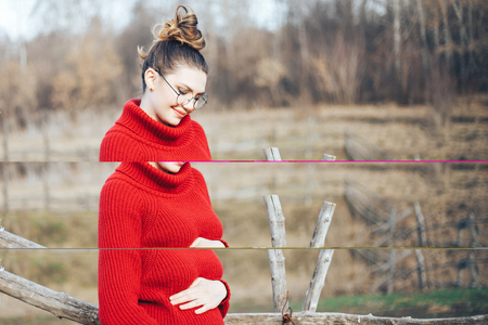 Pregnant woman standing in a field holding his stomach, waiting for the child, summer or spring day, a young mother resting in the open air, in red sweater and round glassesの写真素材