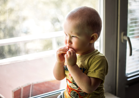 Sad little boy sitting near window and looking outsideの写真素材