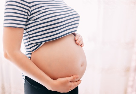 Pregnant woman in t-shirt holds hands on belly on a white background at home, against window. Pregnancy, maternity, preparation and expectation concept. Close-up, copy space, indoors. Beautiful tender mood photo of pregnancy.の写真素材