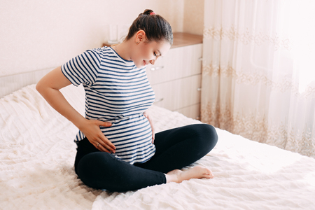 pregnancy, rest, people and expectation concept - happy pregnant woman sitting on bed and touching her belly at homeの写真素材