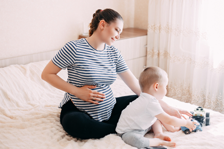 cute blonde little boy hugging a pregnant mother's belly at home, indoor. Mother and son plays in expecting their second child.の写真素材