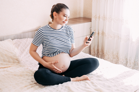 pregnancy, motherhood, technology, people and expectation concept - close up of pregnant woman with smartphone in bed at home, using mobile phone in her bedroomの写真素材