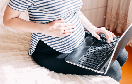 Pregnant woman working on laptop. Cropped image of pregnant businesswoman typing something on laptop while sitting at her working place at homeの写真素材