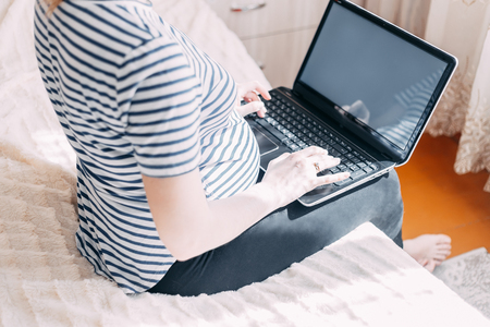 Pregnant woman working on laptop. Cropped image of pregnant businesswoman typing something on laptop while sitting at her working place at homeの写真素材