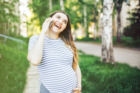 Happy pregnant girl talking on phone at sunset in blooming spring park. pregnant girl with phone lit by rays of sunの写真素材