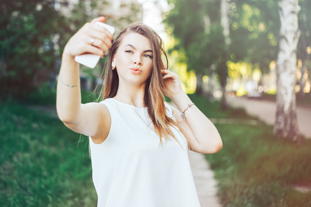 Sexy young woman with long blond hair doing selfie on summer day in park. Cheerful girl making selfie on summer day in park. Close up smiling girl photographing selfie on a mobile phoneの写真素材