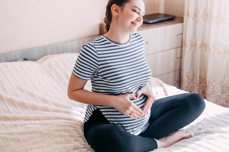Pregnant woman in t-shirt holds hands on belly on a white background at home, against window. Pregnancy, maternity, preparation and expectation concept. Close-up, copy space, indoors. Beautiful tender mood photo of pregnancy.の写真素材