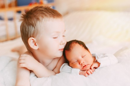Happy little brother with newborn baby lying in a bed together, kissing and hugging. Siblings. New born baby sleeping and two years old brother together. Love. Family.の写真素材