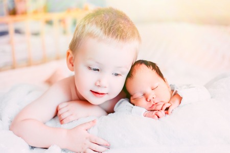 Happy little brother with newborn baby lying in a bed together, kissing and hugging. Siblings. New...