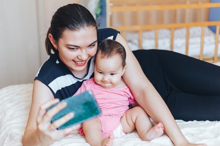 Beautiful young Kazakh mother is making selfie with her adorable little baby and smiling while sitting on sofa at homeの写真素材