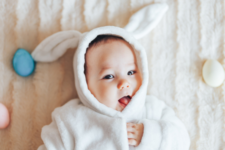 Little child wearing bunny costume on Easter day. baby holding colour eggslying on bed at home, having fun on Easter egg hunt. Happy Easter holiday celebration spring conceptの写真素材