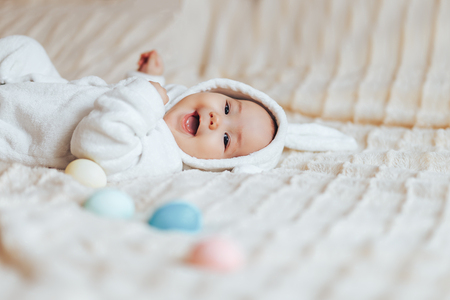 Cute smile happy little baby, playing with colorful easter eggs lying on bed in white rabbit hare costume look at cameraの写真素材