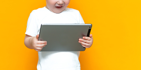 Surprised blonde three years old boy with his mouth open surprised, holding in his hands a tablet pc and looking at the camera on yellow background.の写真素材
