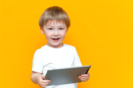 Surprised blonde three years old boy with his mouth open surprised, holding in his hands a tablet pc and looking at the camera on yellow background.の写真素材