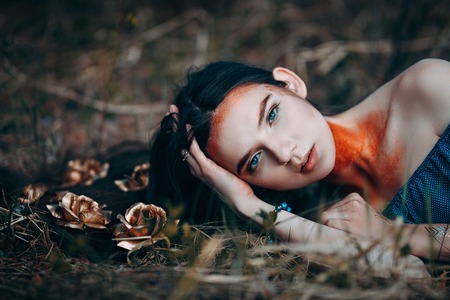 portrait of beautiful brown-haired woman in a blue dress lying on grass, with golden roses on her long hair, in the fairy forest, golden paint shimmer on her neck fantasy conceptの写真素材