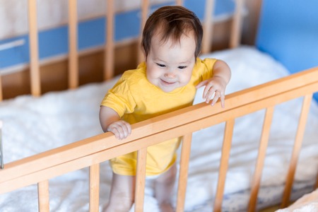 Happy baby girl with dark hair laughing and smiling in yellow t-shirt mixed race kazakh and german standing in cot cribの写真素材