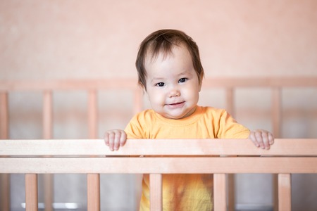 Cute little baby girl with dark hair standing in crib at home in yellow t-shirt.の写真素材