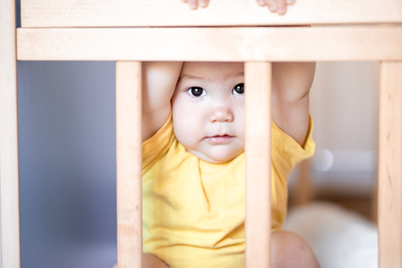 Cute baby sitting in cot looking across the room through the wooden frame. She is afraid of something. baby hide in crib.の写真素材
