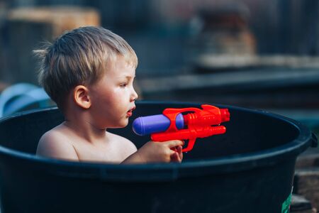 little blonde boy sitting in black pelvis in yard, swim, hot summer have water gun.