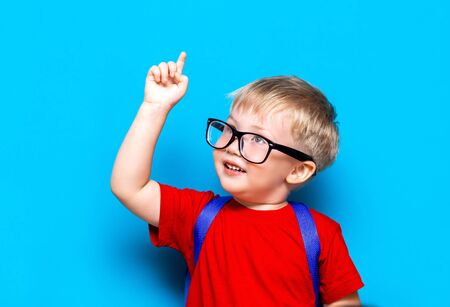 Small boy in red t-shirt. Close up studio photo portrait of smiling boy in glasses with schoolbag and book show something with his finger up, over his head.の写真素材