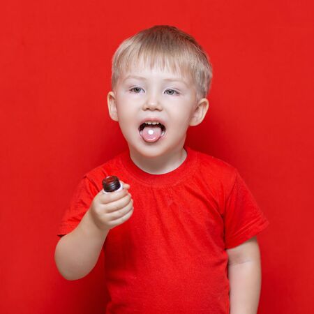 small three years old boy wants to eat pill, tablet on his tongue, can bottle in hand. red background.の写真素材
