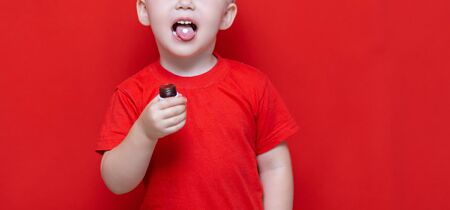 small three years old boy wants to eat pill, tablet on his tongue, can bottle in hand. red background.の写真素材
