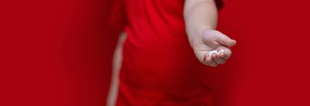 little sad boy kid hold a lot of pills in hands. red t-shirt, red background dangerous concept,...