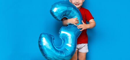 small cute blonde boy on blue background holding foil-coated sphere baloon blue colour. happy birthday three years old.の写真素材