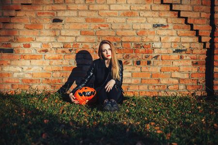 modern Halloween woman. Attractive witch holding pumpkin 'trick or treat'. Woman dressing in leather jacket and black skirt. October halloween. bricks background.の写真素材
