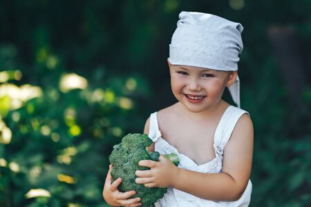 litle happy smiling farmer boy in white overalls and grey hairband holding fresh organic broccoli in hands. garden, harvest season.の写真素材