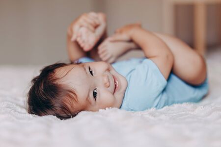 cute laughing one year old girl lying on bed and looking at camera touch her feet.