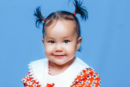 Cute one year old mixed race Asian Caucasian german girl wearing red dress smiles sweetly looking at the camera on blue background.の写真素材