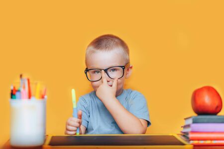 Happy blonde clever boy is sitting at a desk in glasses and smiling. Ready for school. Back to school. Apple, pens and books on desk.の写真素材