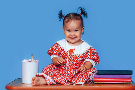 Cute one year old mixed race Asian Caucasian german girl wearing red dress smiles sweetly looking at the camera on blue background.の写真素材