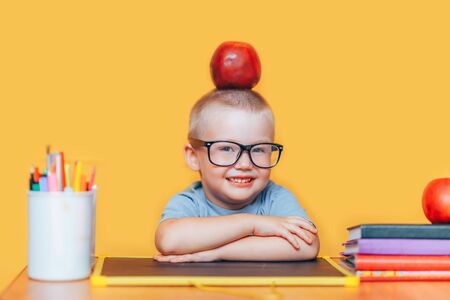 Happy cute clever boy is sitting at desk and smiling. Child is on a yellow background. First time to school. Back to school. pupil in glasses with apple on his head.の写真素材
