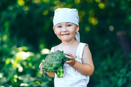 litle happy smiling farmer boy in white overalls and grey hairband holding fresh organic broccoli in hands. garden, harvest season.の写真素材