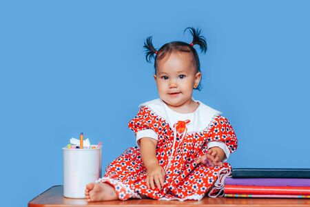 Cute one year old mixed race Asian Caucasian german girl wearing red dress smiles sweetly looking at the camera on blue background.の写真素材
