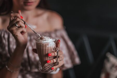 young woman sitting in cafe holding spoon ang glass with coffee or milkshake with whipped cream.の写真素材