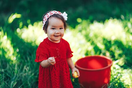 baby girl in red dress eating pear in summer garden near red bucket.の写真素材