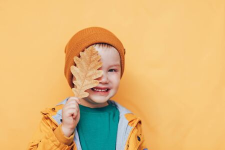 Autumn mood. Happy boy covered eyes with autumn leaf. Autumn time. Smiling baby with yellow leaves in hand. Seasonal fashion. Autumn clothing. kids fashion. Leaf fall. boy in golden clother, orange hat.の写真素材